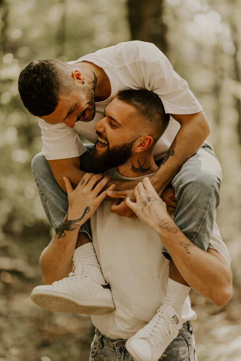 Couple d'hommes s'amusant, l'un portant l'autre sur ses épaules, lors d'un shooting photo couple en forêt.