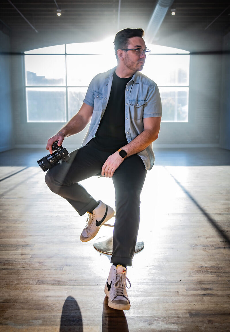 Alex Atkinson in a thoughtful pose on a stool, camera in hand, within an expansive and empty studio setting.