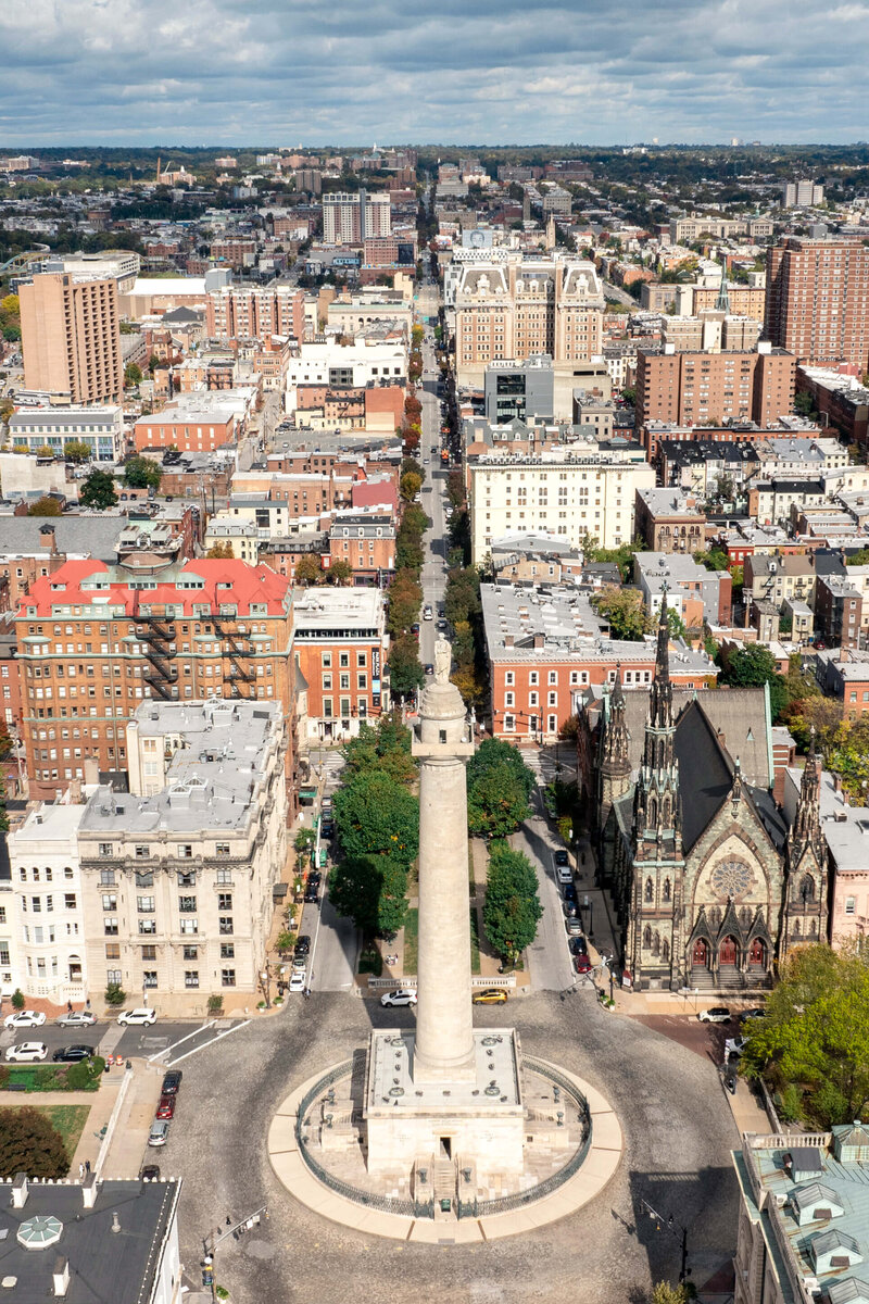 Aerial view of Baltimores Washington Monument surrounded by urban buildings and streets. A large church with intricate architecture stands nearby. The cityscape extends into the distance under a partly cloudy sky.