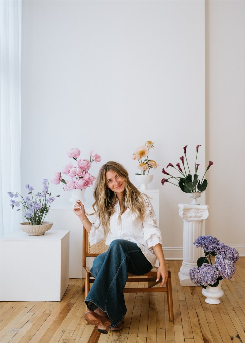 Saratoga Springs floral designer, Annalee, sitting in a wood and cane chair surrounded by modern, simplistic floral arrangements