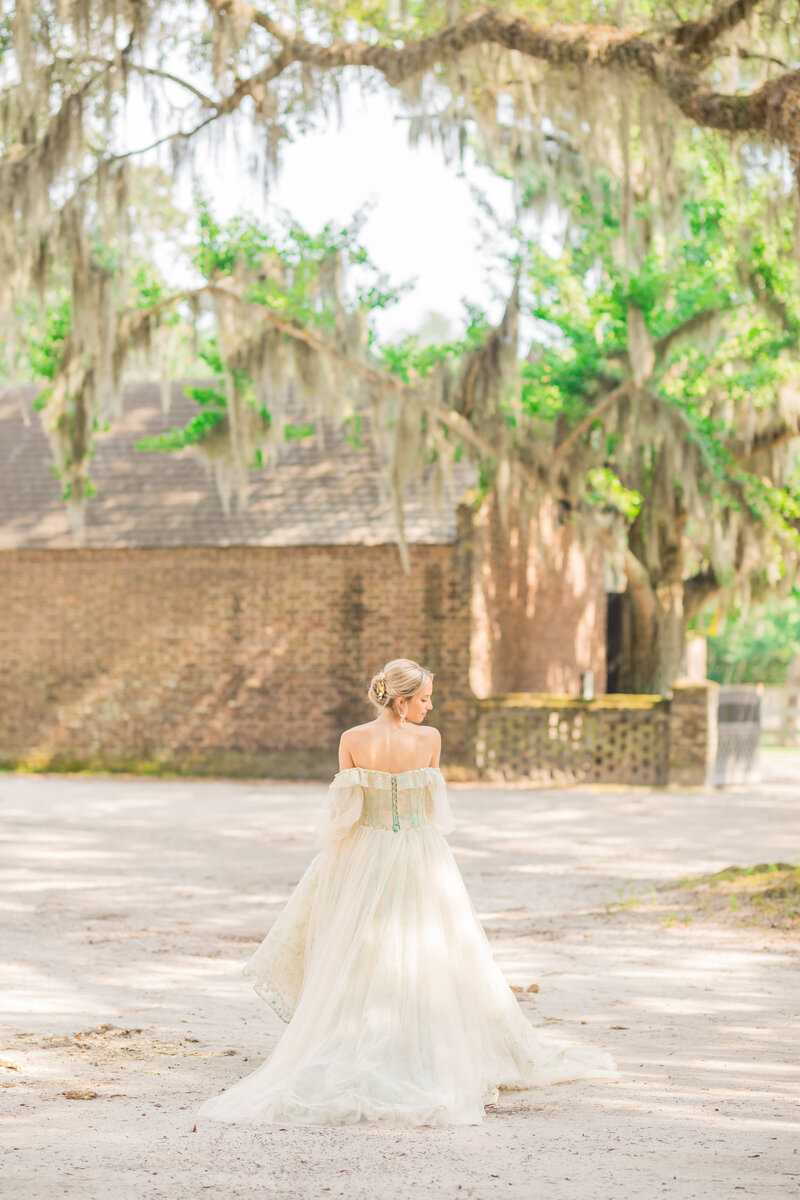 A bride in an off-the-shoulder gown walks gracefully beneath Spanish moss at a historic brick building in Middleton Place, Charleston.