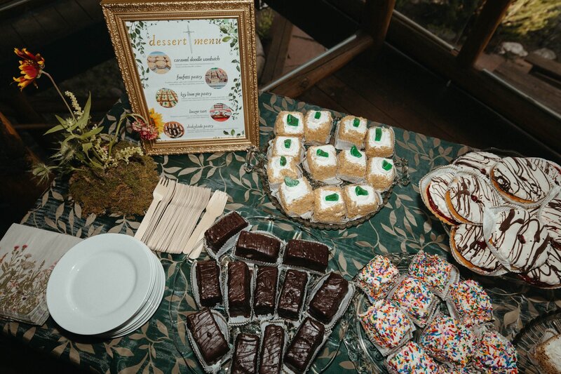 A wedding dessert display with multiple platters full of decorated mini desserts, plus a menu in a gold frame and small mossy floral arrangement