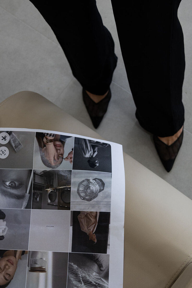 Close-up of a woman in a sharp black pant suit and black pointy-toed heels, looking down at a printed image board placed on a beige chair.