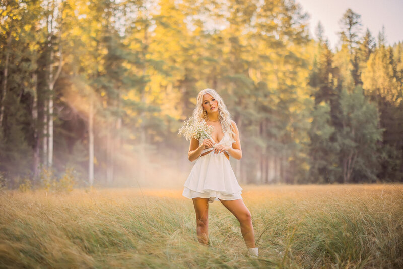 senior girl with long blonde wavy hair wearing a hot pink mini dress sitting in a golden field with blue glacier national park mountains in the background at sunset