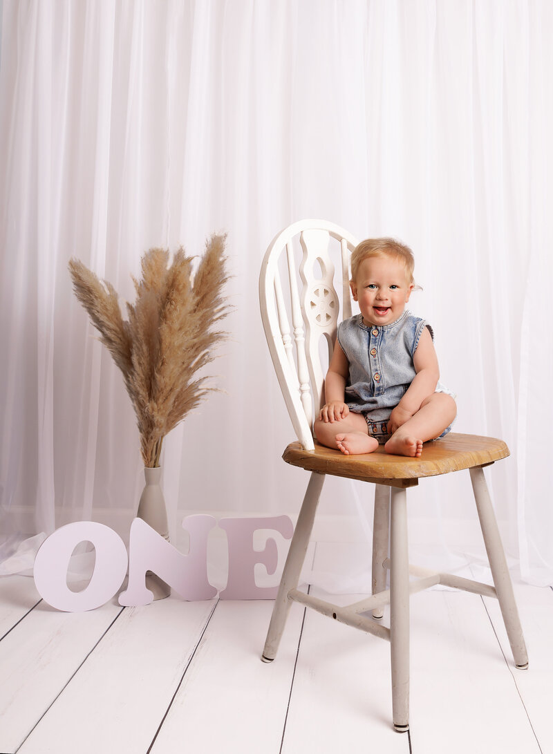 Brooklyn first birthday photoshoot. Baby boy is wearing a denim romper and sitting on a chair for a studio first birthday photoshoot. He is smiling at the camera. Captured by best Brooklyn, NY first birthday milestone photographer Chaya Bornstein.