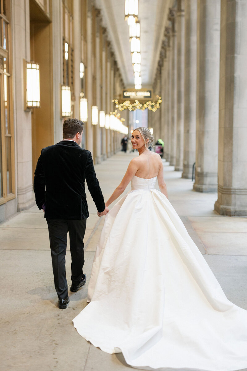 A bride in a stunning white ball gown with a long train and a groom in a black velvet suit walk through a grand corridor with ornate columns and string lights, sharing a loving glance.