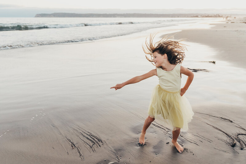 A girl in a yellow dress dances during her family photography session  on a San Diego beach while her hair flips in the air