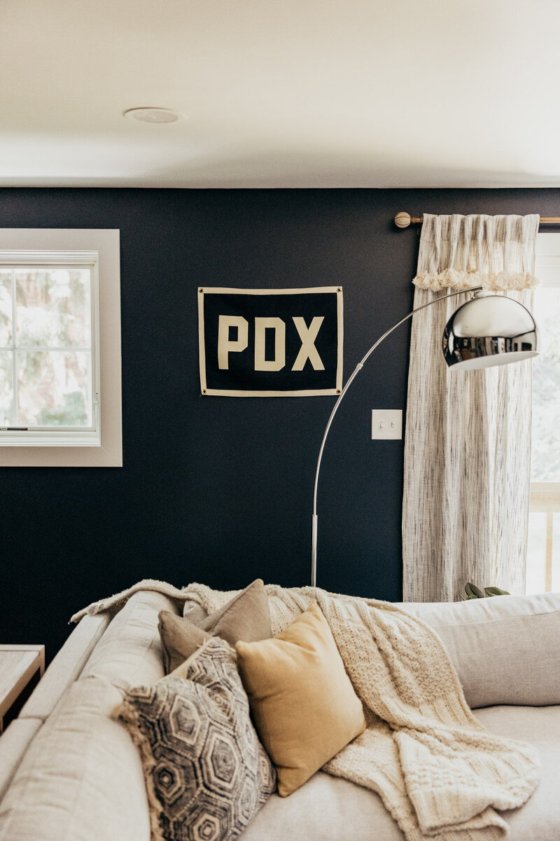A colorful interior design featuring a oversized couch with throw pillows and blanket, dark blue walls with a PDX sign and a modern lamp.