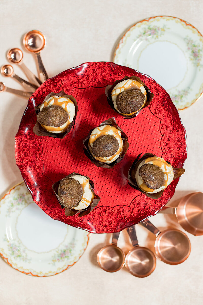 Gourmet cupcakes with caramel drizzle and a spiced cookie topping displayed on a red embossed cake stand. Vintage floral plates and copper measuring spoons surround the dessert, adding a rustic, artisanal touch. Small business branding photography in Columbia, SC, showcasing handcrafted bakery goods and culinary creativity.