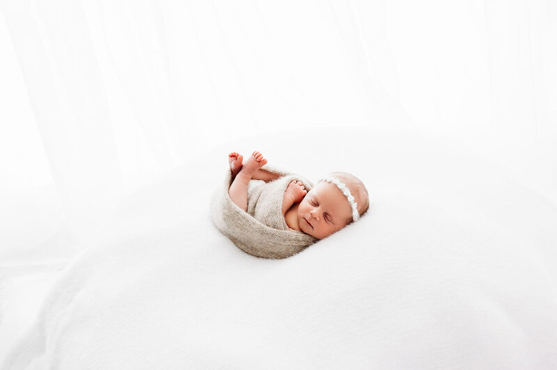 Newborn baby girl swaddled in beige wrap, wearing white headband laying on white blanket