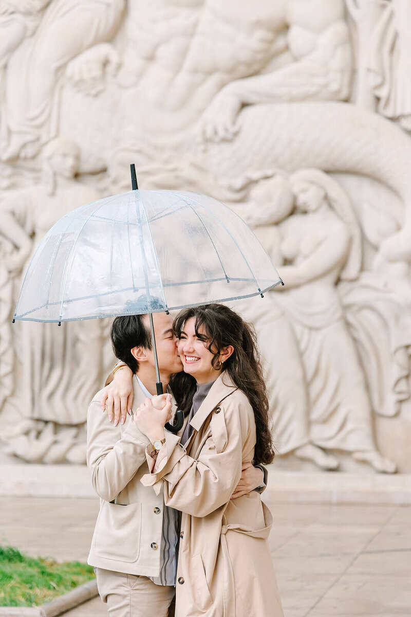Man kissing smiling woman holding an umbrella and wearing a trench coat during a romantic Paris proposal near the Eiffel Tower at Passerelle Debilly and Palais de Tokyo.