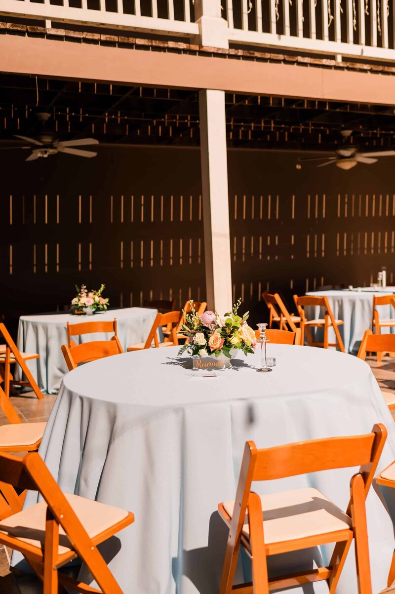 Round tables with light blue tablecloths and wooden chairs are set up outdoors. Each table has a floral centerpiece and place cards. Sunlight creates warm shadows on the scene.
