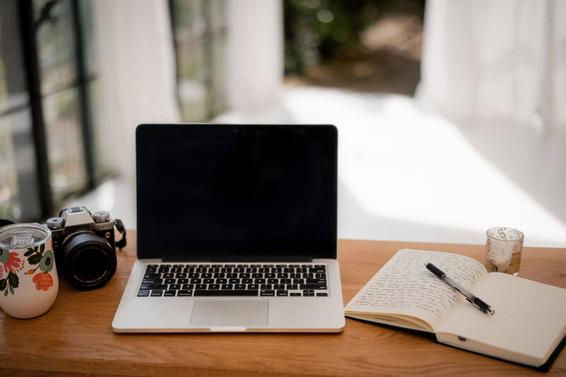 Laptop, Notizbuch, Kamera und Kaffeetasse auf Holzschreibtisch im Tageslicht – Arbeitsplatz von Meg Hoechsmann.