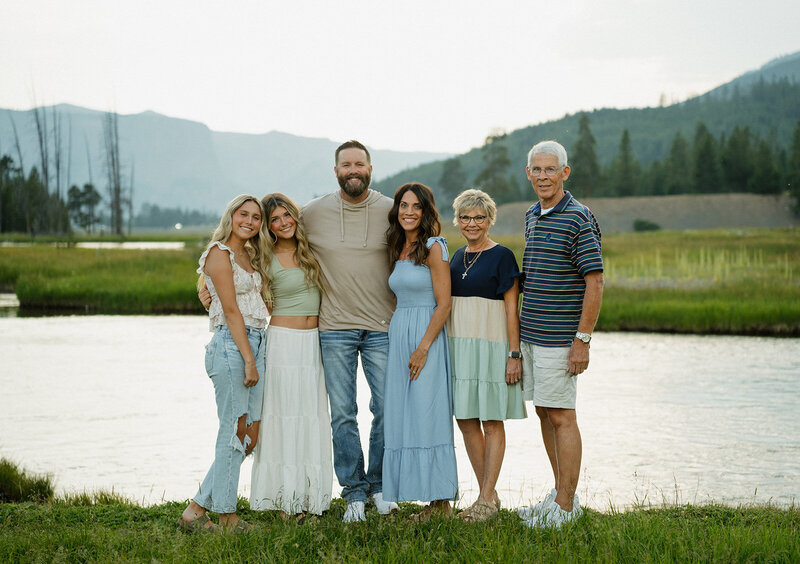 Family photographer in Rexburg Idaho, Adriana Montoya Photo. A posed family photo with a mom, dad, grandma, grandpa, and 2 daughters all smiling at the camera. Behind them is a lake and behind that are trees and mountains.