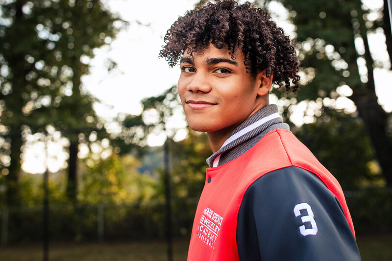 A dark skinned senior boy smiles towards the camera wearing his basketball jacket from a Pittsburgh area school.