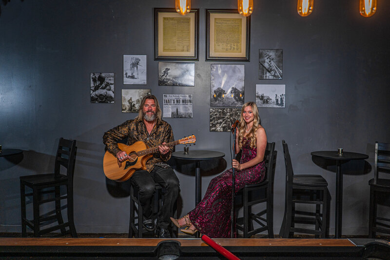 Two people sitting at a hightop table while one holds a guitar and the other has a mic in front of them 