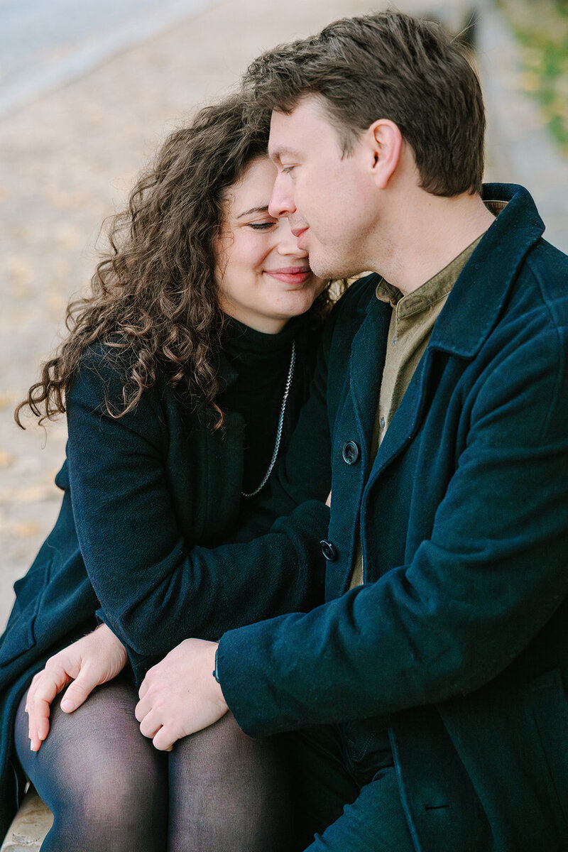 Man and woman sitting closely together near Notre-Dame and Pont Neuf, with the woman leaning into him as he rests his hand on her thigh during a romantic Paris proposal photoshoot.