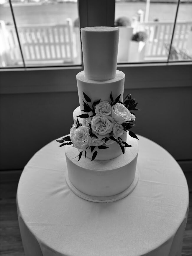A black and white photo of a four tier wedding cake with a cluster of fresh roses and greenery arranged at the center.