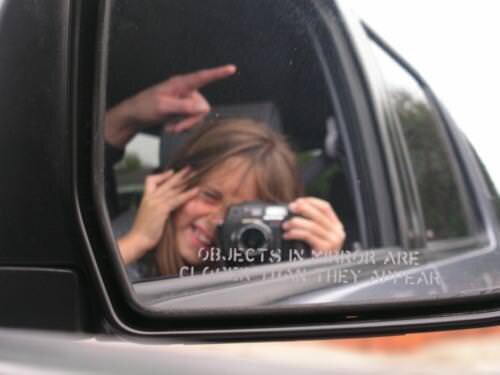picture of a kid holding a film camera in the car, looking through the viewfinder and taking a picture of themself in the rearview mirror. The driver of the car is pointing out of the car. Mirror says "object in mirror are closer than they appear"
