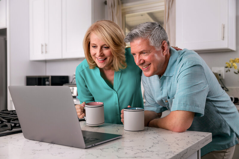 Middle aged husband and wife look at laptop screen and smile during virtual video consult while in their kitchen holding coffee mugs