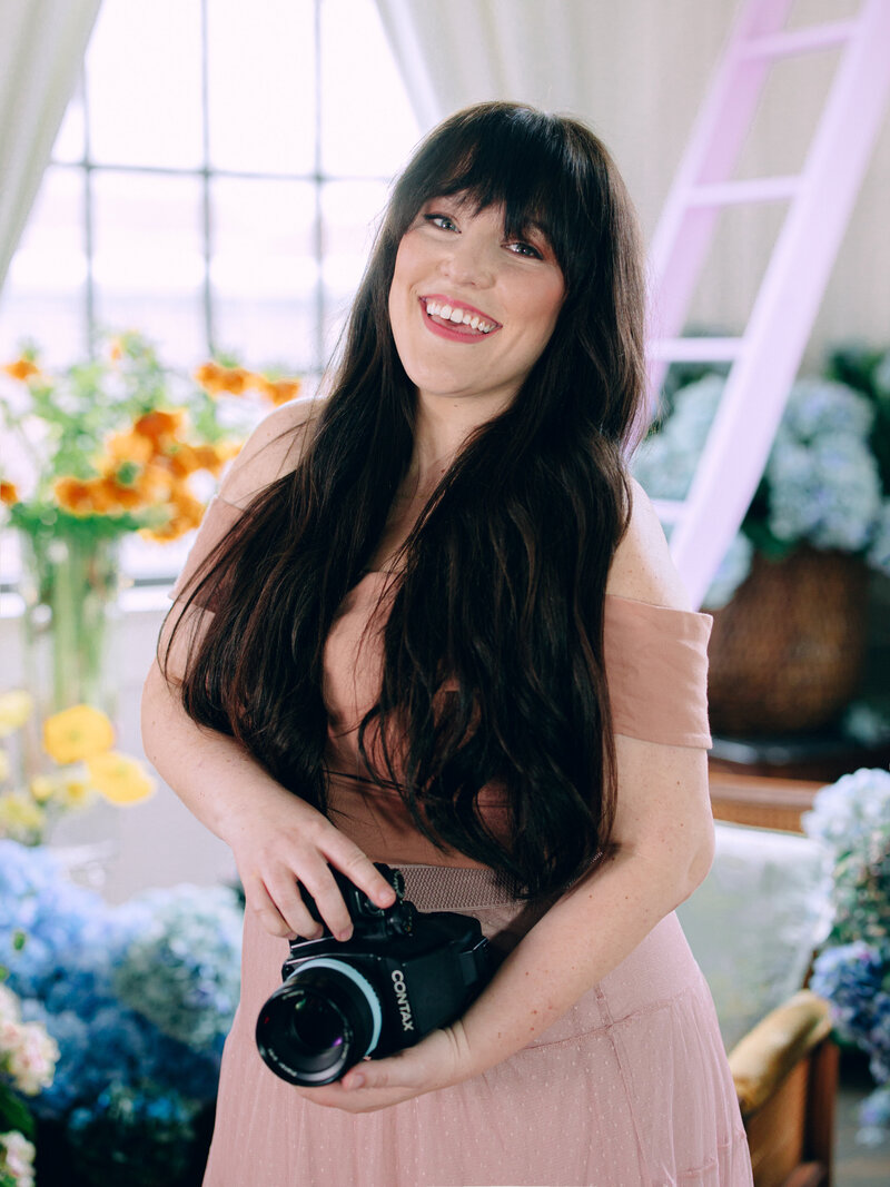 headshot of a woman holding a film camera surrounded by buckets of flowers