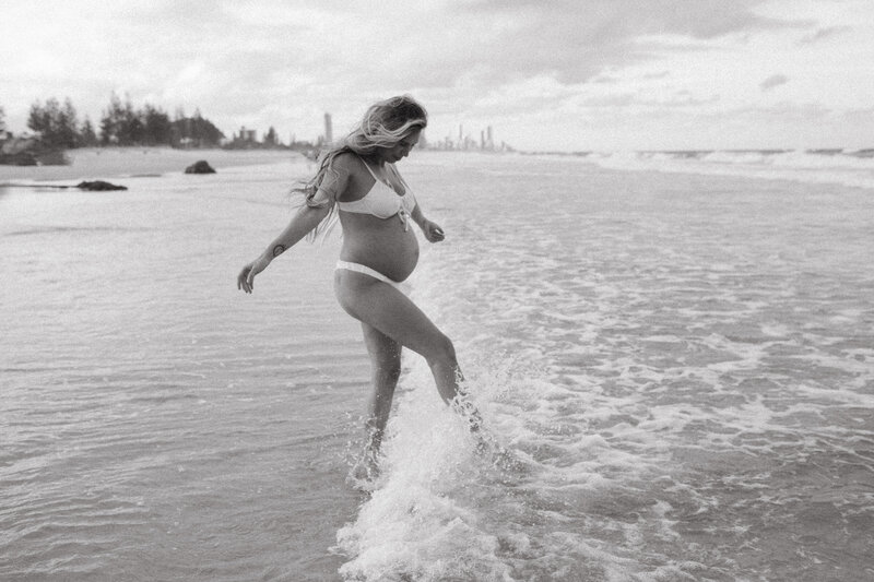 A pregnant mother spalashing in the waves at Burleigh BEach , Surfers paradise cityscape is in the background. It is a candid playful moment