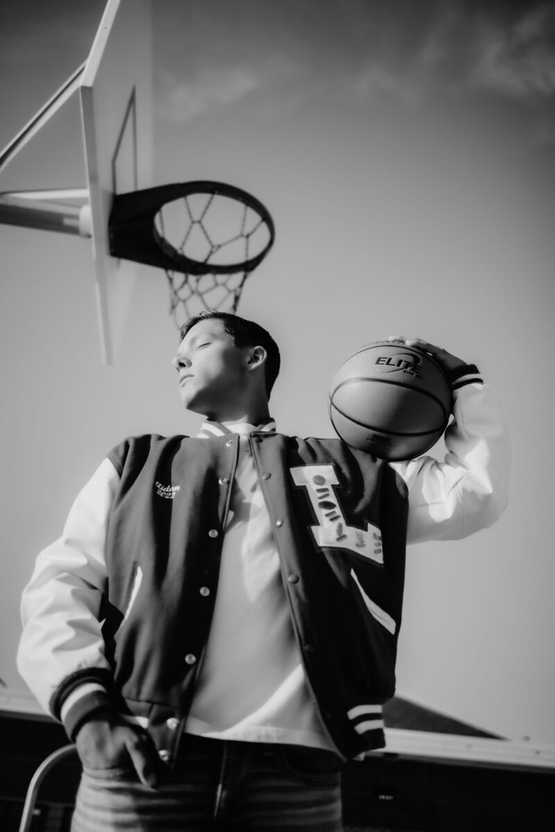 A senior wearing their letterman jacket with a large L on it standing beneath a basketball hoop with a basketball on shoulder during their Senior photoshoot captured by Infinite Productions