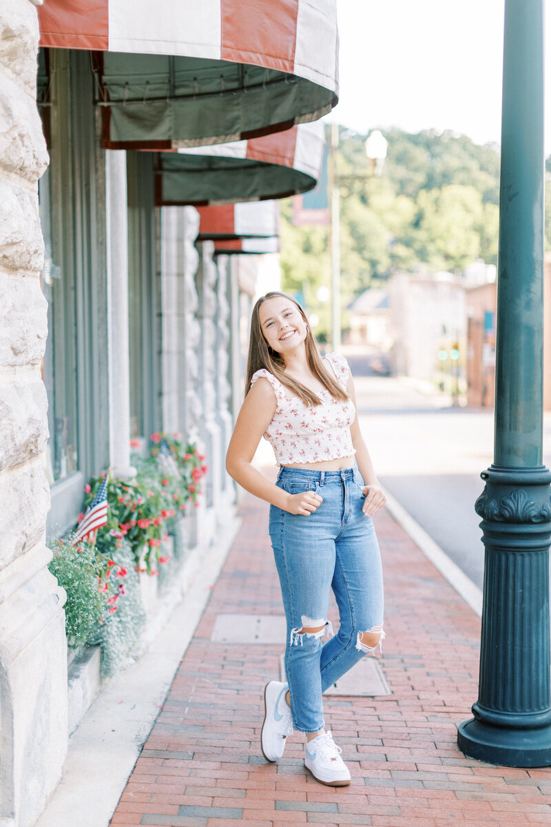 Cute brunette senior girl takes her photos in downtown Staunton, Virginia. She is wearing jeans, and white sneakers with a floral top.