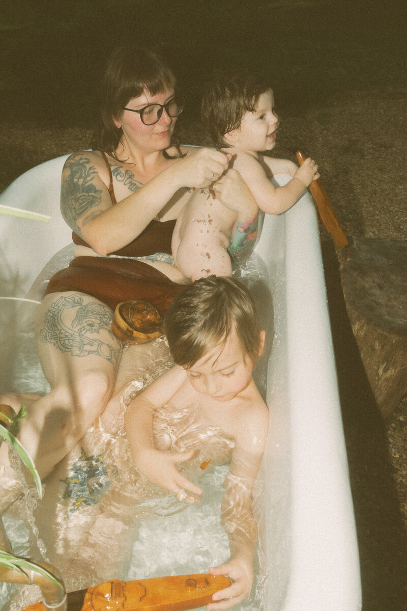 A mother in a cute bathing suit sits in the bath with her two young boys. 