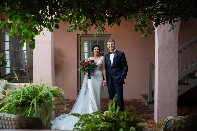 A bride in an elegant white gown with lace bodice and flowing train stands with her groom in a navy blue tuxedo with burgundy bow tie in a charming courtyard at the White Room in historic downtown St. Augustine. They're positioned in front of coral-colored stucco walls under natural greenery and vines, with the bride holding a colorful bouquet of red, purple, and blue flowers. Lush ferns and brick pathways complete the romantic garden setting.