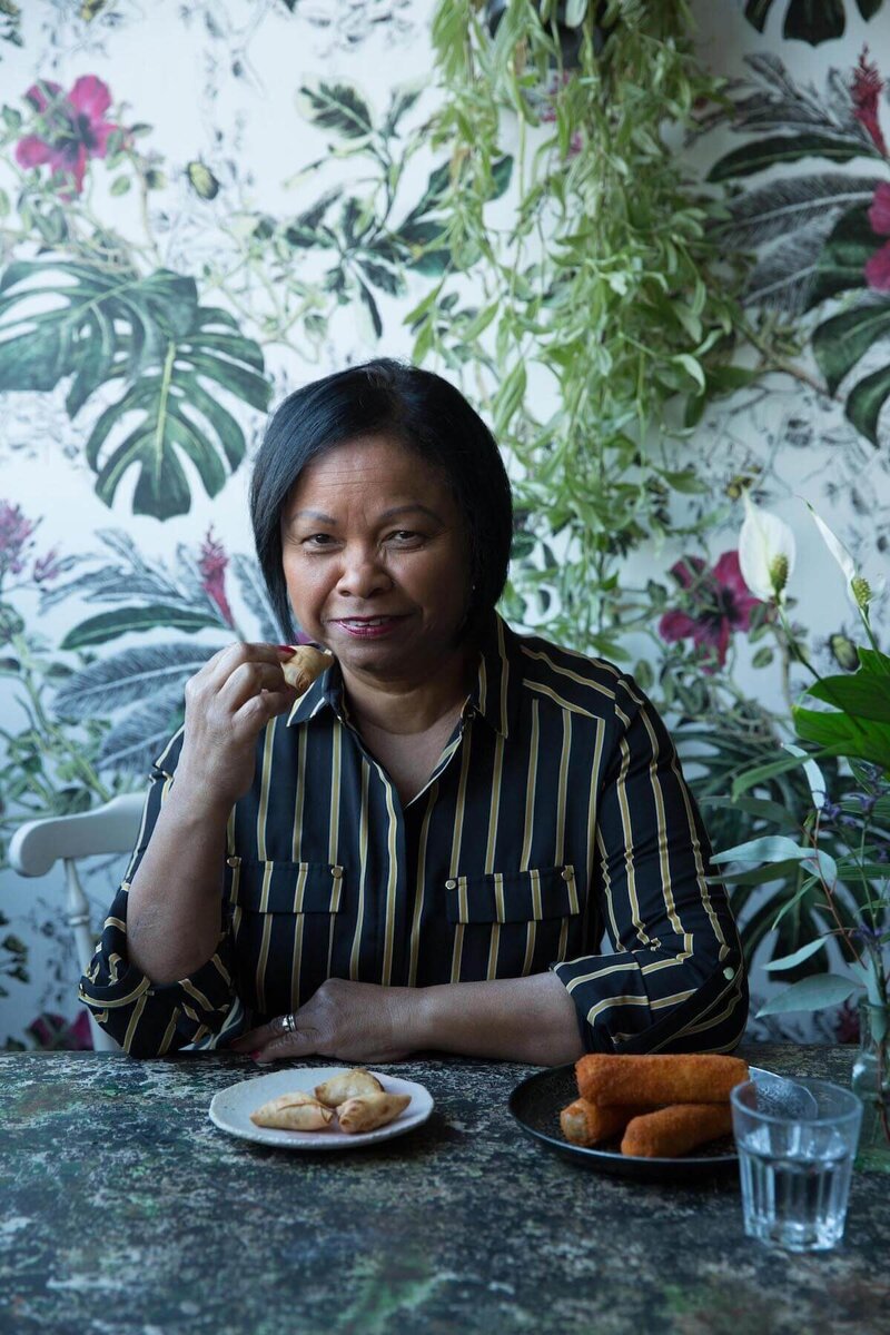 Image of Lilette in a black stripy shirt sitting at a table eating a somosa
