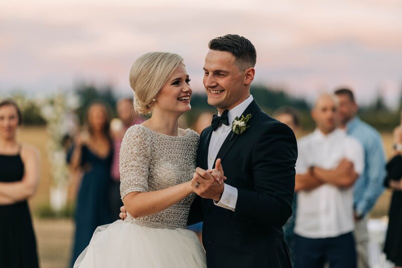 bride and groom share a first dance with the pink sunset sky in the background while their friends look on