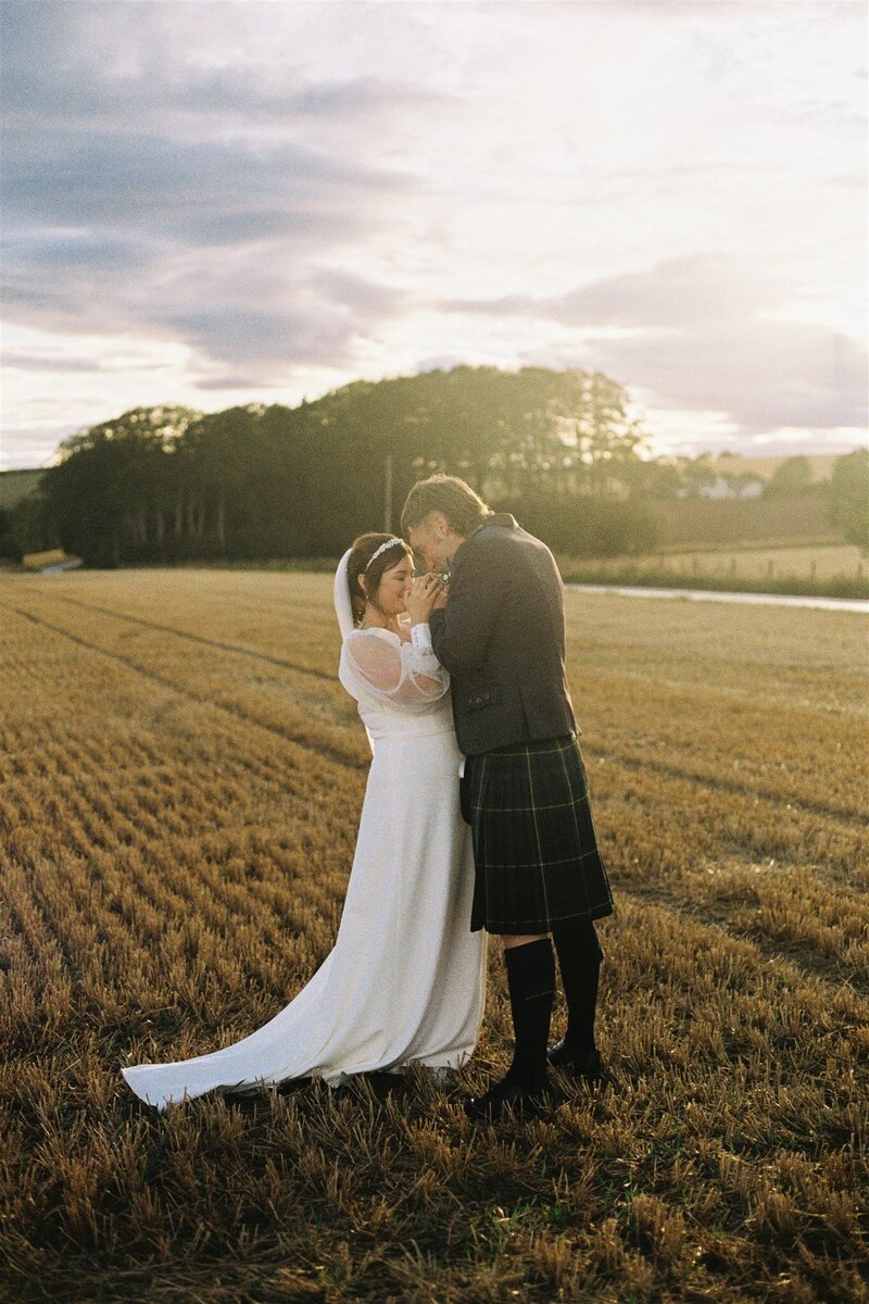 A couple stand in a field chest to chest. They snuggle their hands together and are photographed under a golden hour sky on 35mm film.