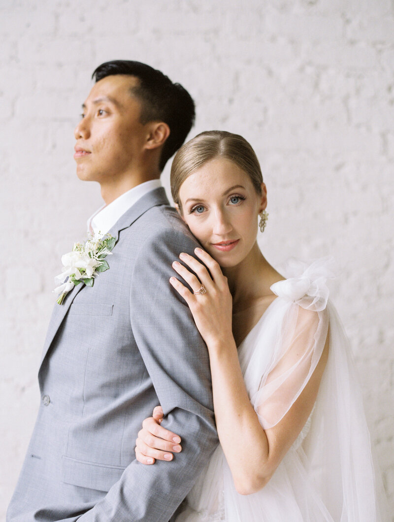 a groom in a light grey suit looking off into the distance and his bride behind him holding on to his arm and resting her head on his back while she is looking at the camera