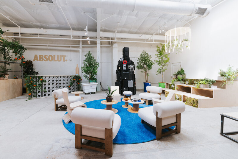 Modern lounge setup for an Absolut event at Tales of the Cocktail, featuring white armchairs around a blue circular rug, wooden tables with plants, greenery throughout the space, and a large black sound system installation against the wall.