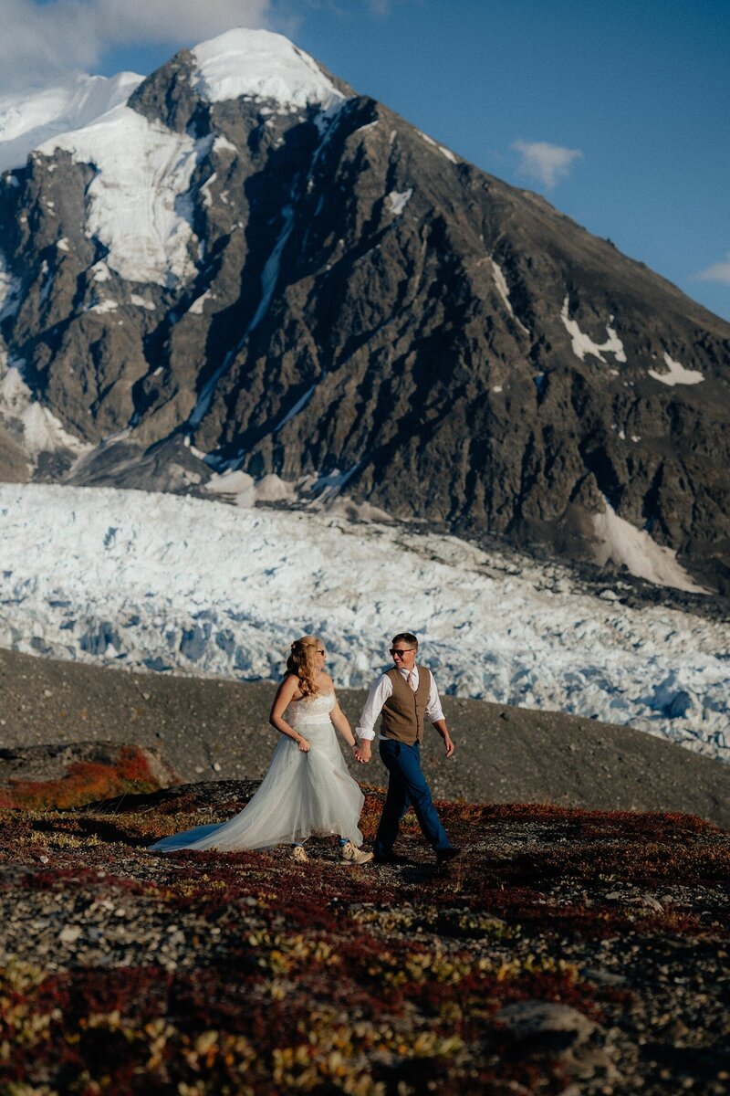 Bride and groom holding hands on Knik Glacier with helicopter in the background during adventurous Alaska elopement.