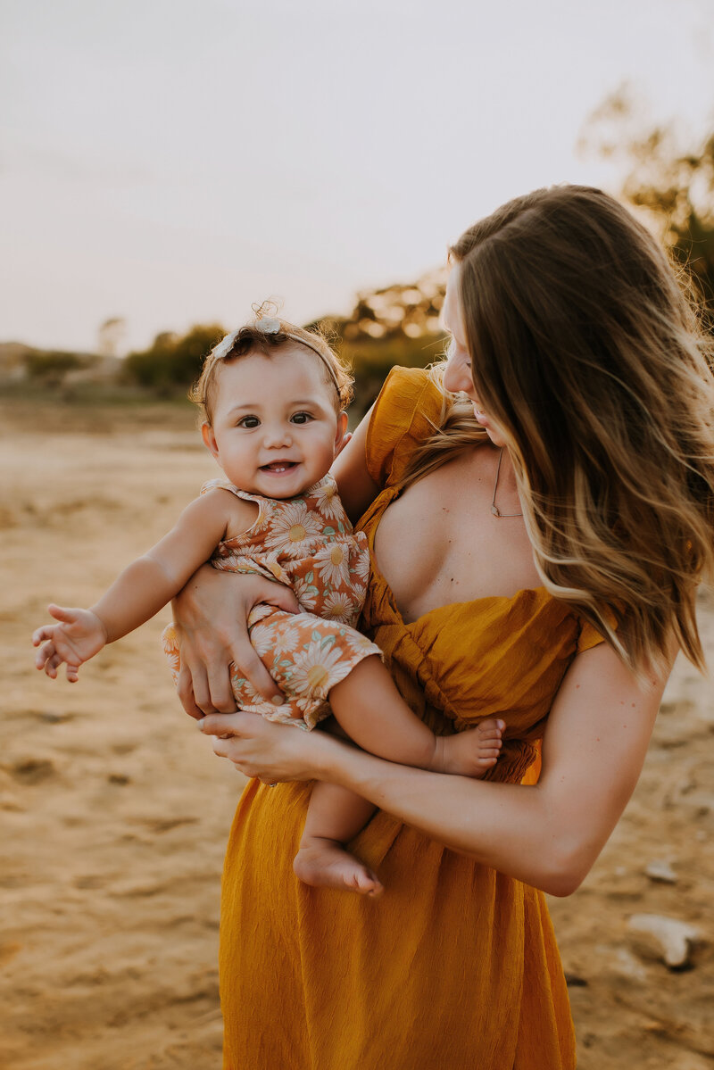 A mother in a linen mustard dress holding her baby girl in the sand at Murrell Park in Flower Mound, TX