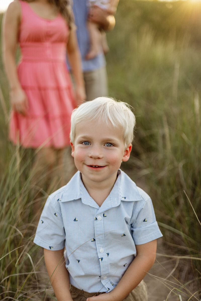  A young boy stands in a grassy field in West Michigan, smiling softly as the golden-hour light casts a warm glow. A timeless childhood portrait capturing innocence and joy.