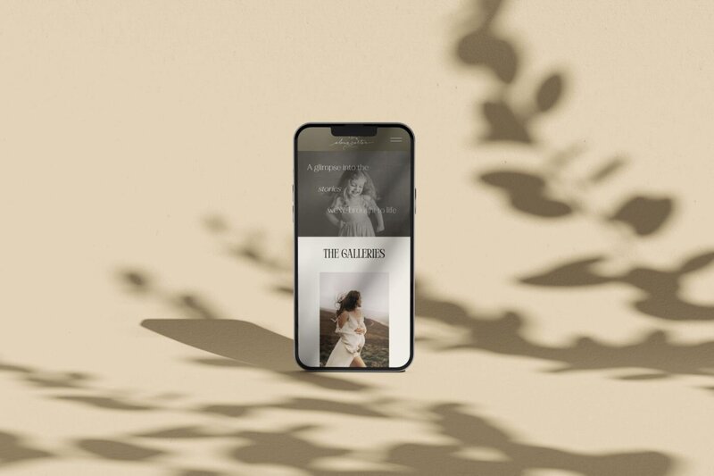 Smartphone standing upright on a beige surface with soft leaf shadows, displaying a photography website gallery page with a black-and-white child portrait and a woman in a flowing dress outdoors.
