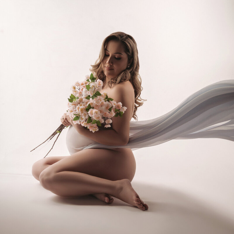 CT maternity photographer captures beautiful pregnant mother sitting on the floor wrapped in a flowing light blue fabric while holding a bouquet of small pink flowers in front o a white backdrop