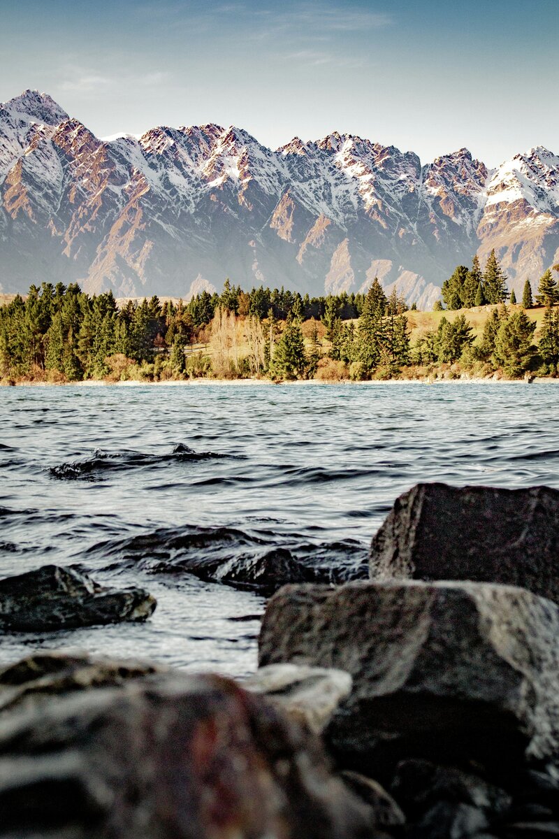 Rocky shoreline with gentle waves on Lake Whakatipu in Queenstown, surrounded by evergreen trees and autumn foliage, with dramatic snow-capped mountains in the background under a clear sky
