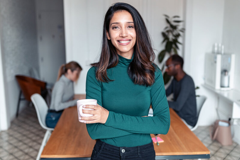A Latina woman has dark hair and is wearing a green turtleneck with a cup of coffee in her hand. She is smiling.