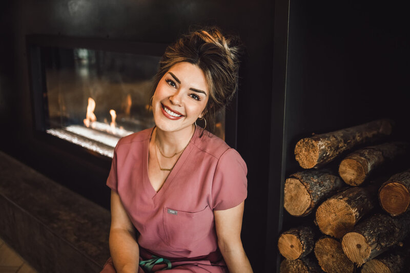woman sitting in front of logs and fireplace wearing pink scrubs while smiling