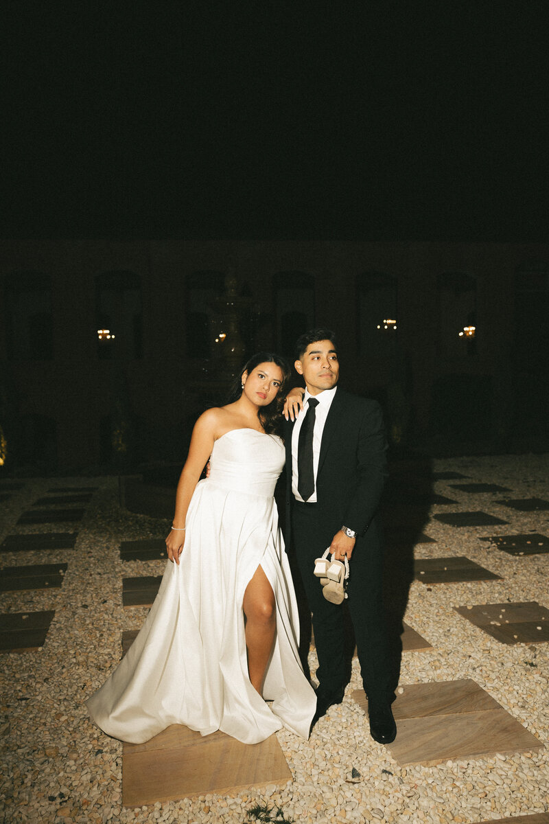 Newlyweds lean together on a stone patio at night while groom holds bride's shoes