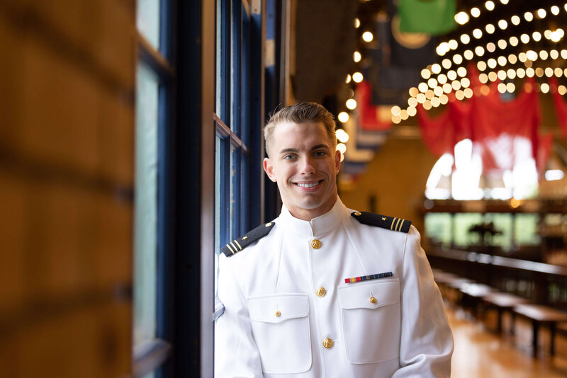 Midshipman in whites smiling and leaning on window with golden lights and colorful flags of Dhalgren Hall in background
