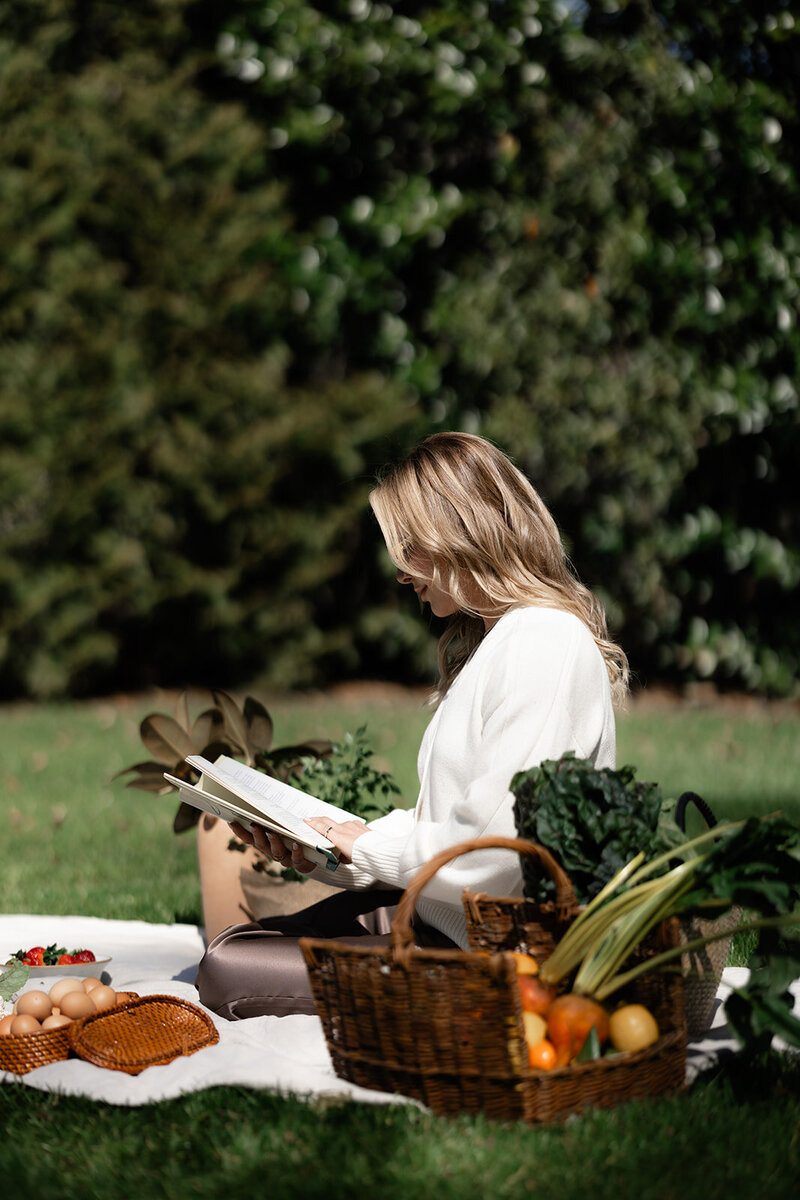 Tracy Greenstein sitting on a picnic blanket reading a nutrition book beside a basket of healthy food and vegetables