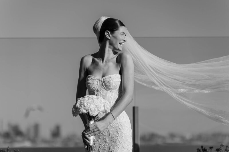 Black and white portrait of a bride with her veil blowing in the wind above a city skyline