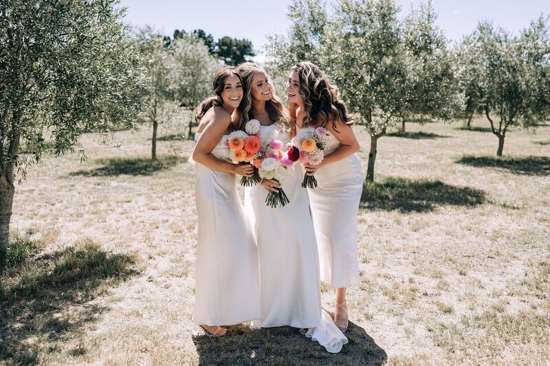 bride in white dress and bridesmaids in ivory to match with pink and orange dahlia bouquets in waipara olive grove