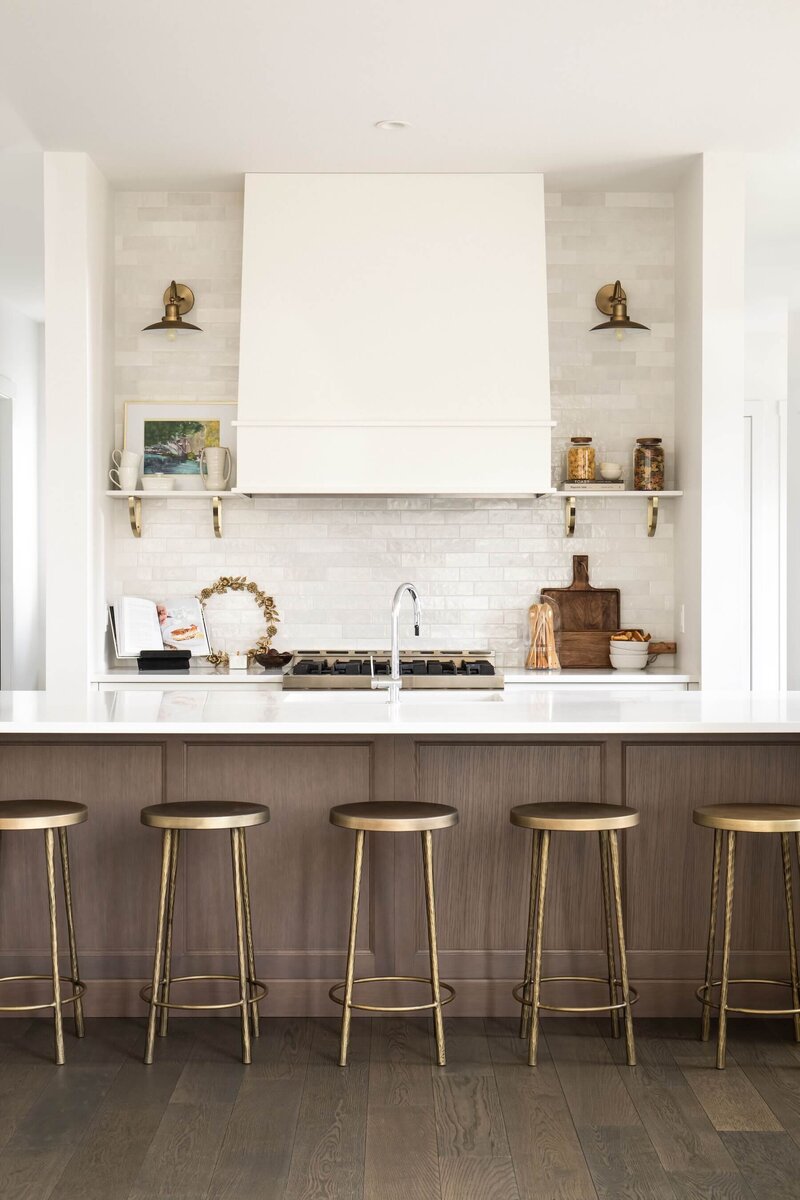 A gorgeous kitchen with a wooden hood fan, open shelving, and brass counter stools. 