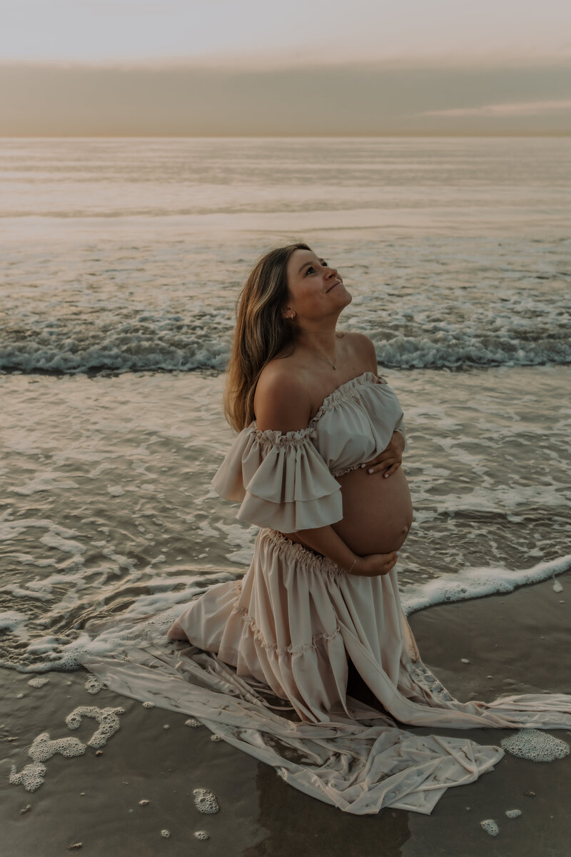 Zwangere vrouw in sierlijke jurk poseert in het water op het strand tijdens zwangerschapsshoot in Knokke.